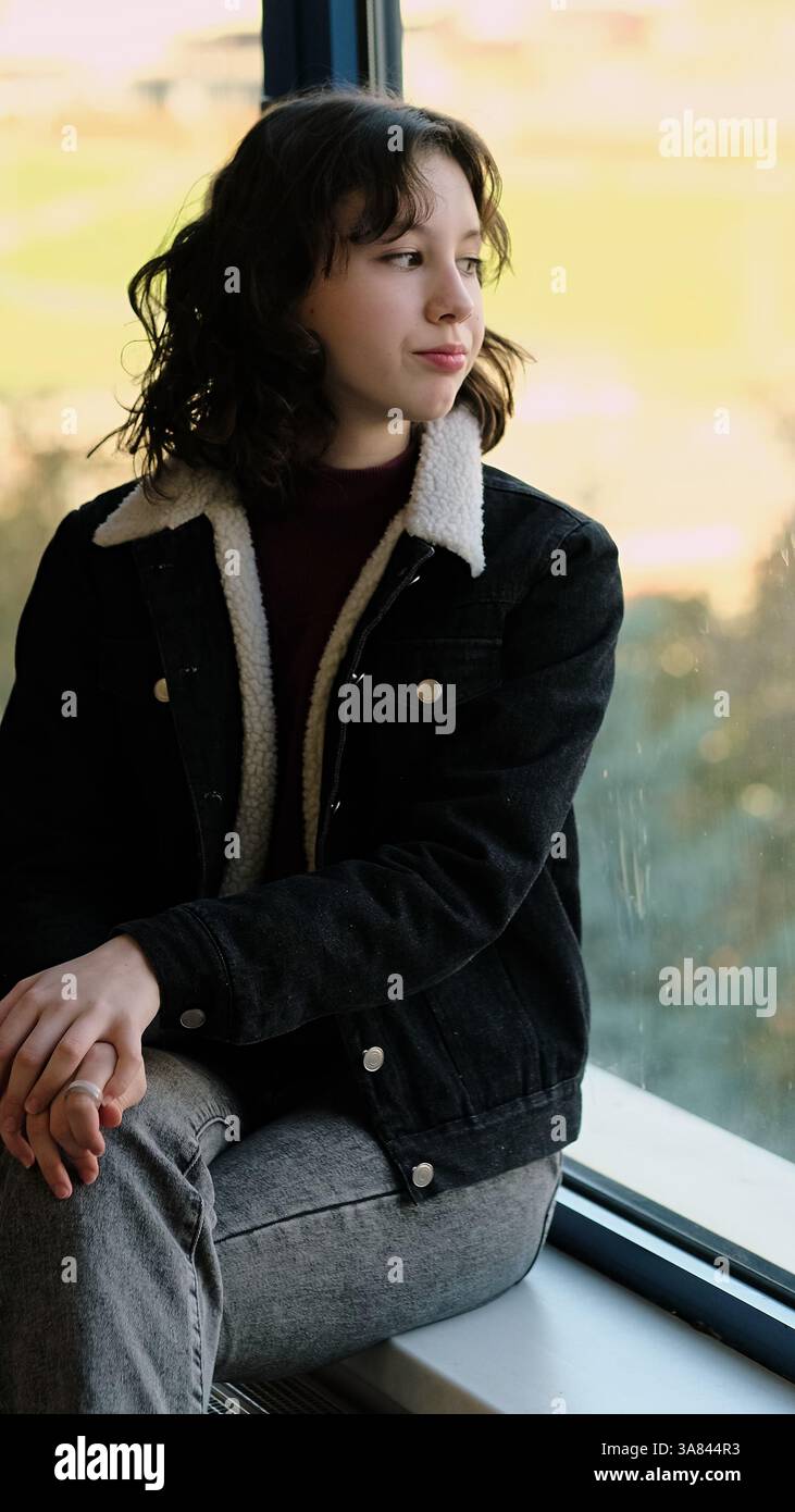 Ein nachdenkliches Mädchen sitzt leise an einem Fenster, ihre Haltung ruhig und introspektiv. Das Foto fängt die ruhige Schönheit persönlicher Momente und des selbst ein Stockfoto