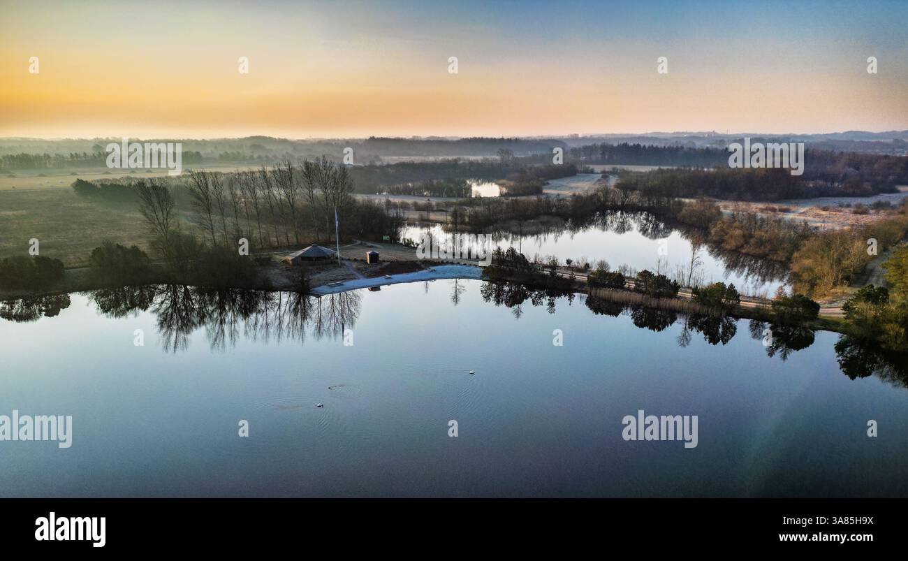 Eine atemberaubende Luftaufnahme eines ruhigen Sees, der den lebendigen Himmel bei Sonnenaufgang reflektiert, umgeben von frostiger Wintervegetation und ruhiger Natur, Show Stockfoto