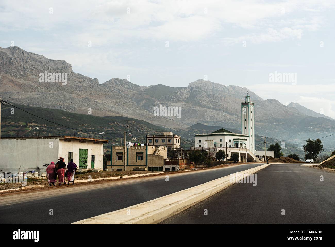 Ländliche Straße durch ein marokkanisches Dorf mit einer Moschee und hoch aufragenden Bergen in der Ferne, Marokko Stockfoto