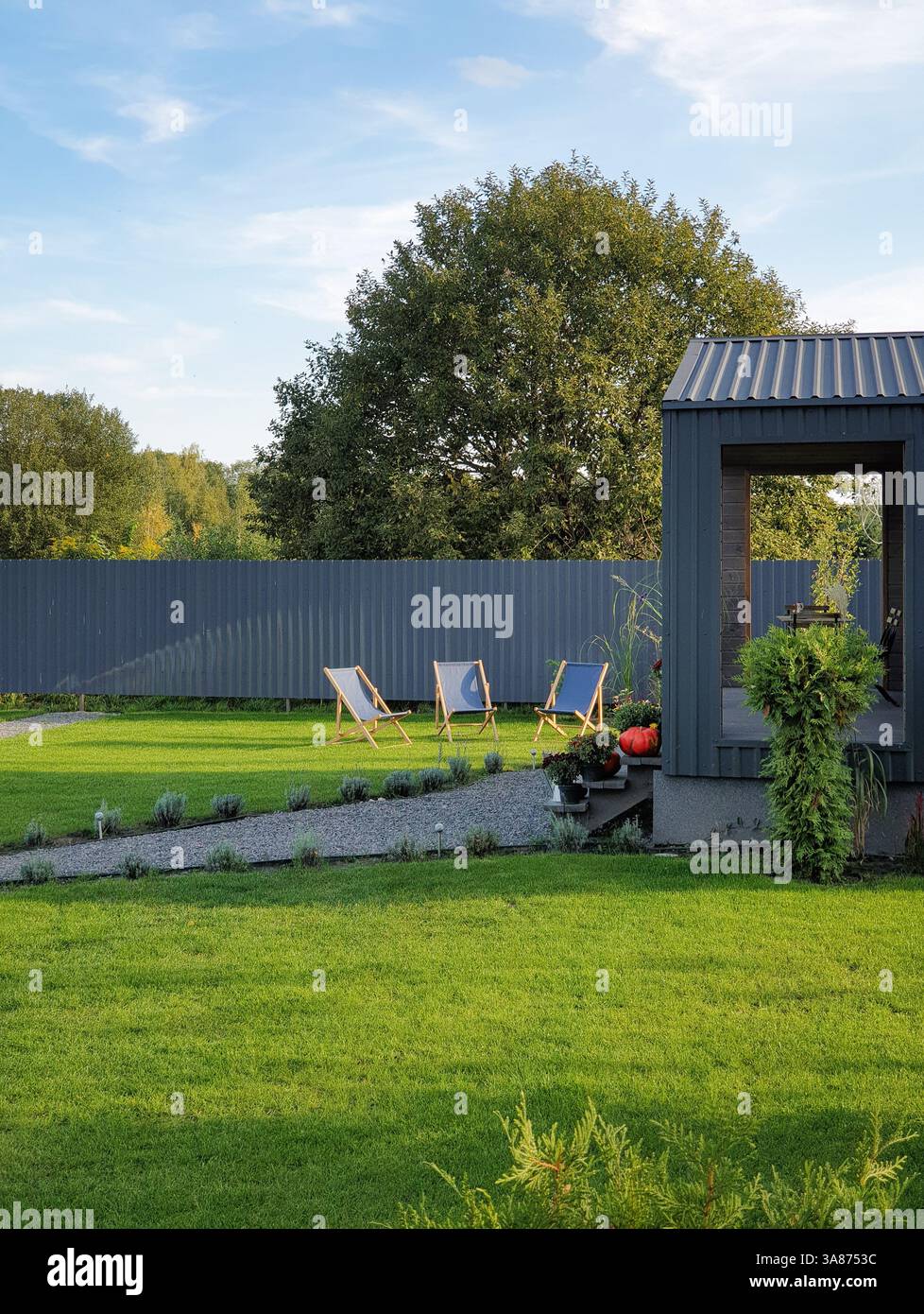 Hinterhof in Sonnenstrahlen am Nachmittag: Drei Liegestühle auf einem Rasen, Veranda mit Sommerhaus und Fußweg. Mehrere Bäume hinter dem grauen Zaun. Stockfoto