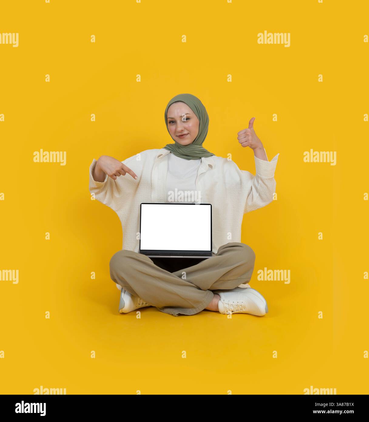 Muslimische Frau im Hijab sitzt auf der Etage und empfehle die Webseite. Zeigen Sie den Laptop mit leerem Bildschirm nach oben, Daumen nach oben Geste. Großartiges Angebot, befürworten Sie die Konzeptidee. Stockfoto