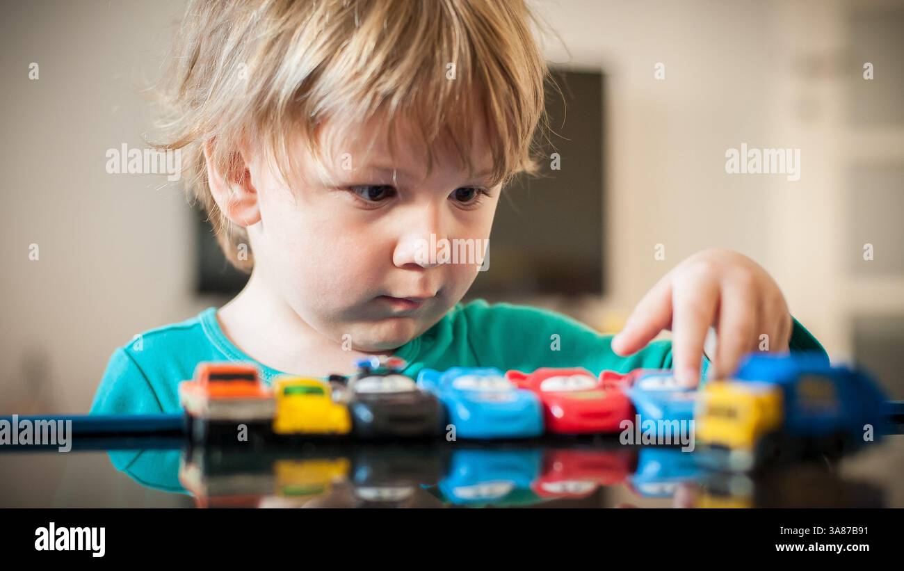Ein kleines Kind untersucht und organisiert eine Sammlung von Spielzeugautos auf einer reflektierenden Oberfläche und zeigt Neugier und Konzentration in einem hellen setti Stockfoto
