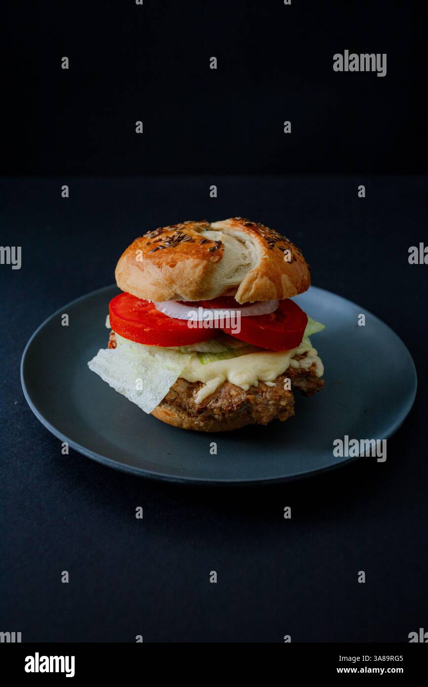 Beef Burger mit Gurke, Tomate und Salat auf einem schwarzen Teller mit dunkler Essthetik und stimmungsvollem Hintergrund, im vertikalen Format erfasst Stockfoto