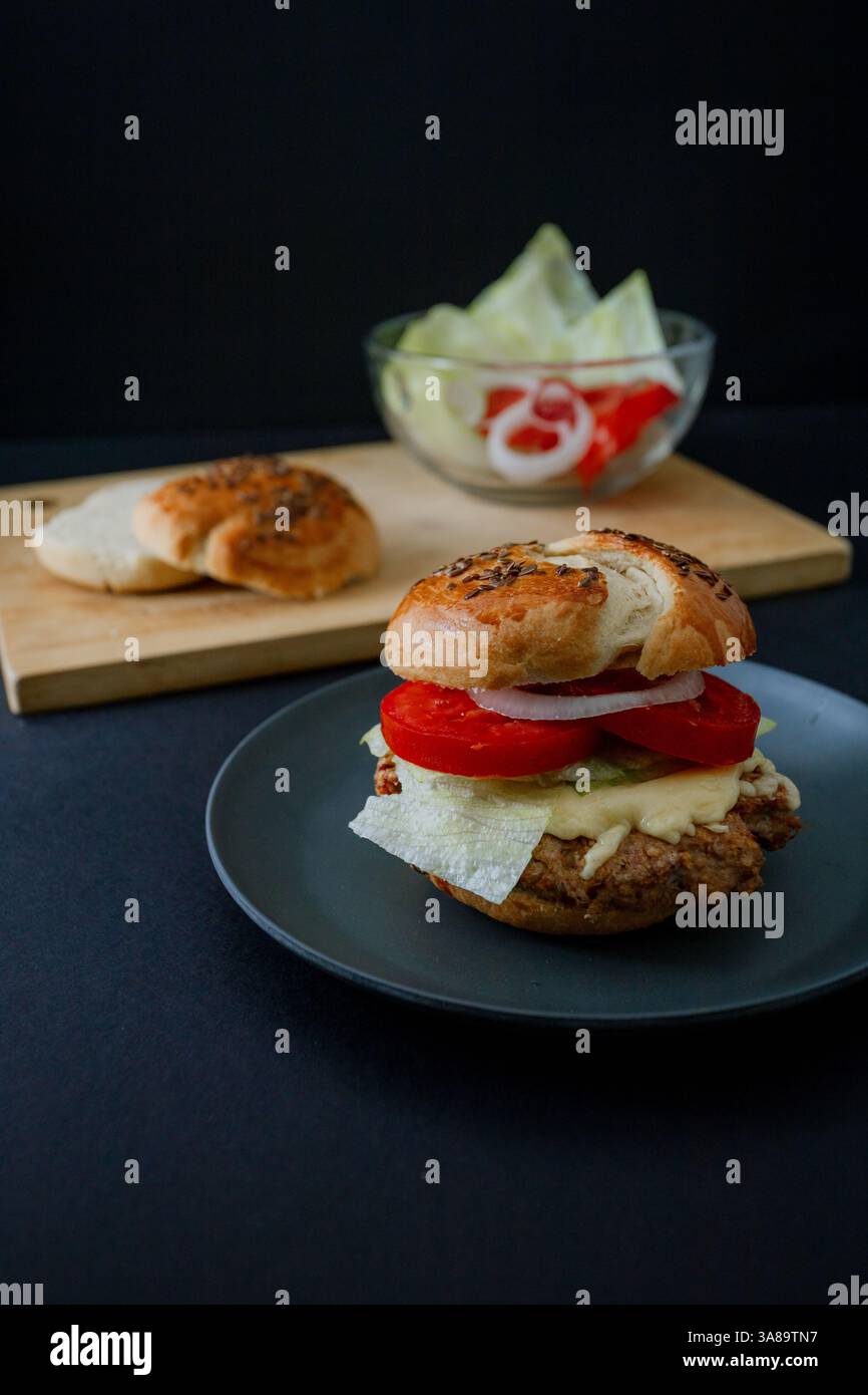 Beef Burger mit Gurke, Tomate und Salat auf einem schwarzen Teller mit dunkler Essthetik und stimmungsvollem Hintergrund, im vertikalen Format erfasst Stockfoto