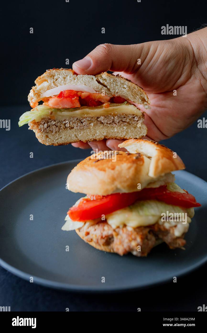 Beef Burger mit Gurke, Tomate und Salat auf einem schwarzen Teller, von einer Hand gehalten, mit dunkler Essthetik und einem stimmungsvollen Hintergrund Stockfoto