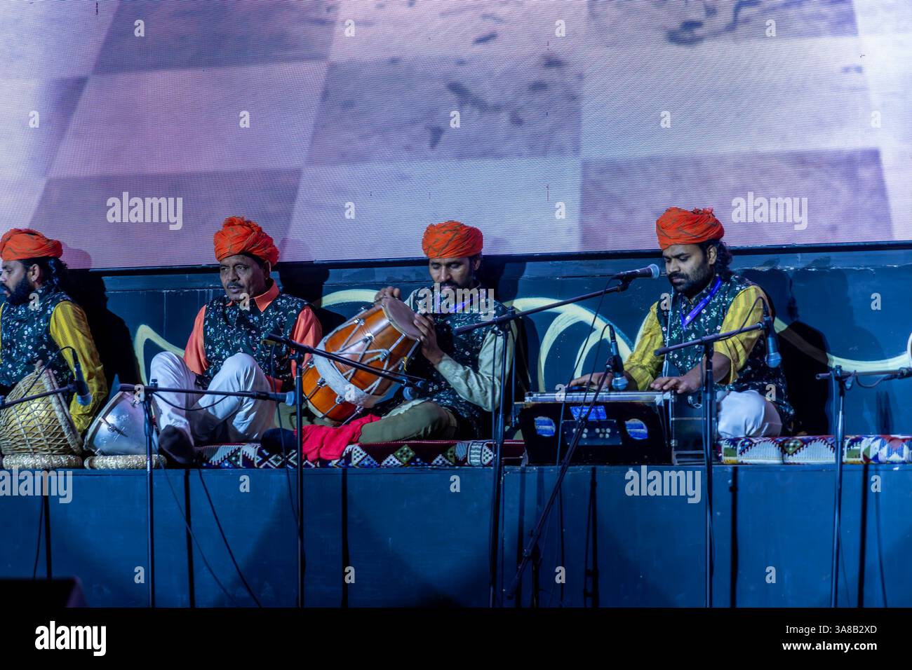 Indische Musiker treten in traditionellen Outfits mit orangen Turbanen und gemusterten Westen aus der indischen Kultur auf und spielen mit Instrumenten auf der Bühne. Stockfoto