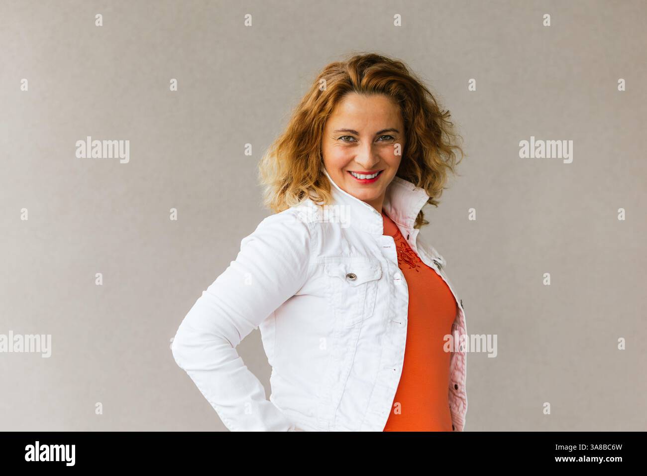 Frau mittleren Alters in einem orangefarbenen Oberteil und einer weißen Jacke, die selbstbewusst lächelt, Hände auf den Hüften trägt, vor einem neutralen Hintergrund steht und Selbstbewusstsein ausstrahlt Stockfoto