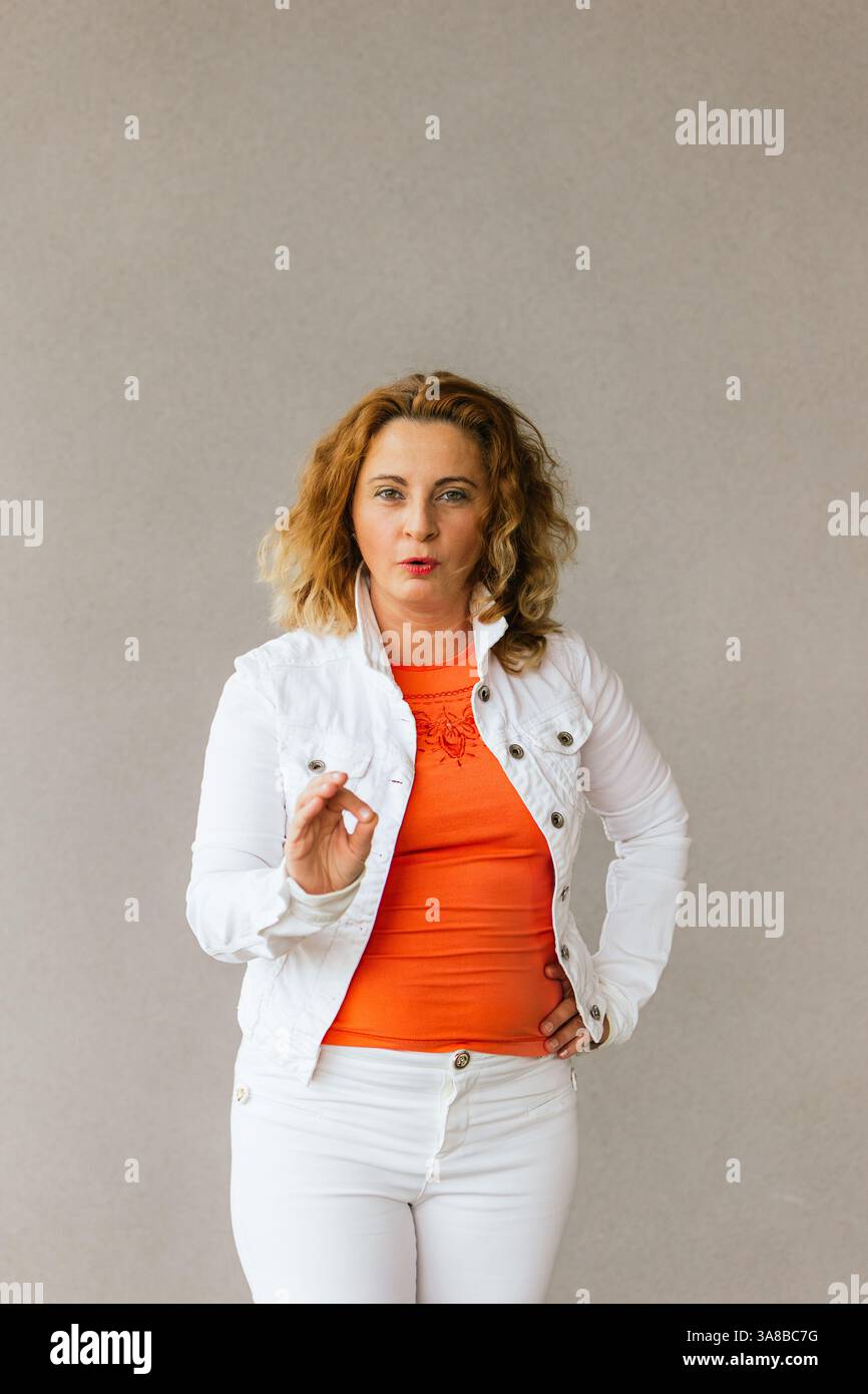 Frau mittleren Alters in orangefarbenem Oberteil und weißer Jacke, die eine ausdrucksstarke Handbewegung macht und mit einem ernsten und selbstbewussten Look vor einem neutralen Rücken steht Stockfoto Frau mittleren Alters in orangefarbenem Oberteil und weißer Jacke, die eine ausdrucksstarke Handbewegung macht und mit einem ernsten und selbstbewussten Look vor einem neutralen Rücken steht Stockfoto