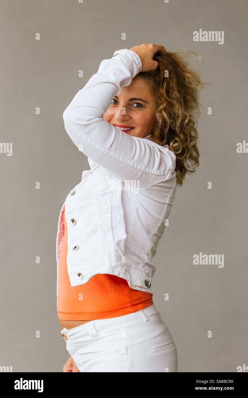 Lächelnde Frau mittleren Alters in orangefarbenem Oberteil und weißer Jacke, die sich spielerisch mit der Hand im Haar posiert und mit einem Vertrauten vor einem neutralen Hintergrund steht Stockfoto