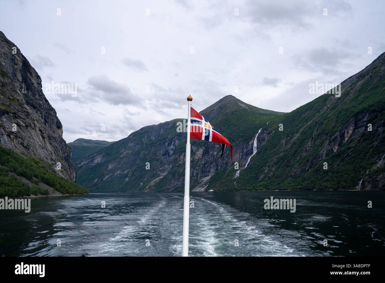 Blick vom Heck eines Bootes, das durch den Geirangerfjord in Norwegen segelt, mit der norwegischen Flagge und einer spektakulären Landschaft aus Bergen und Stockfoto