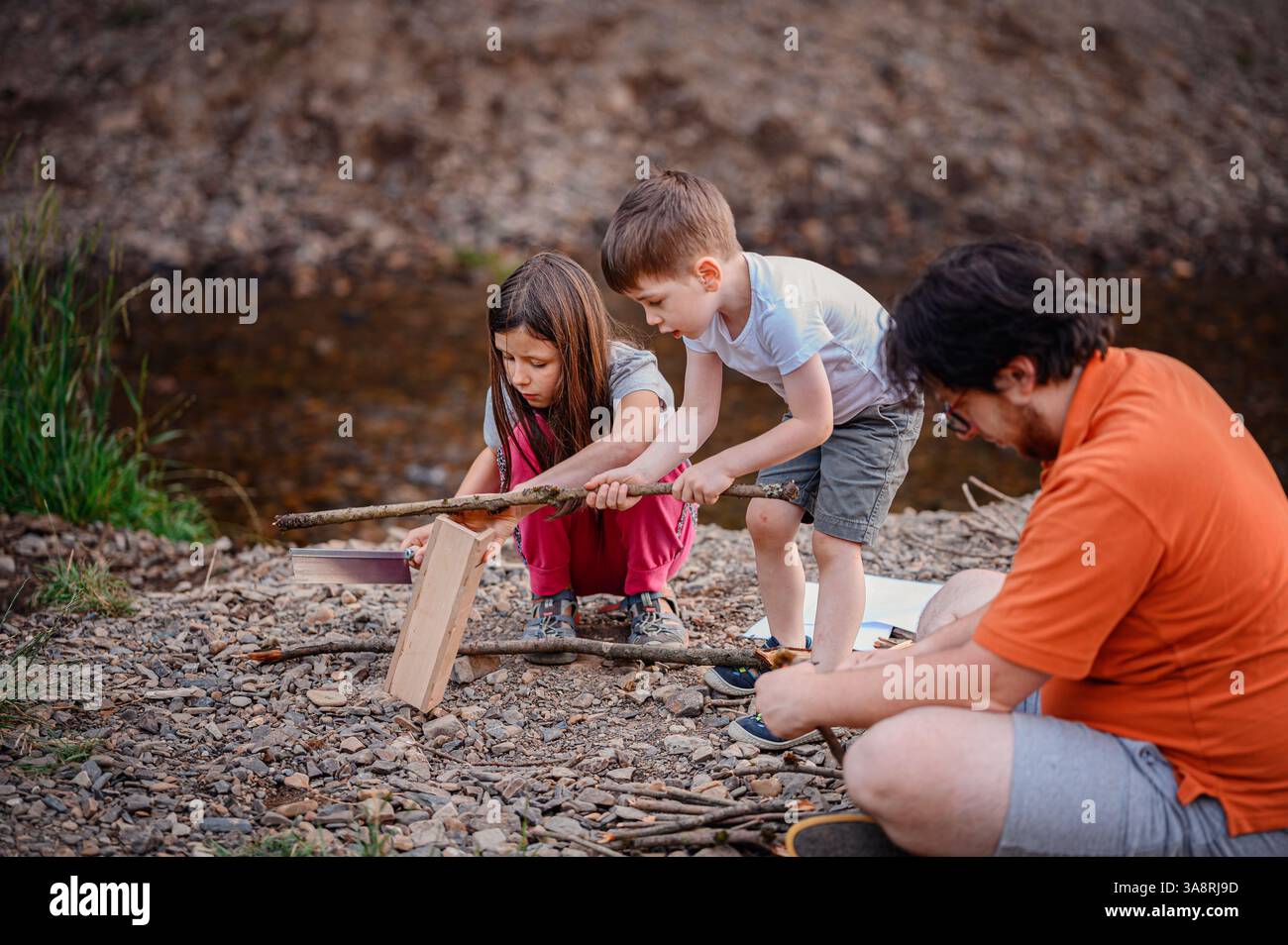 Ein Junge und ein Mädchen arbeiten zusammen, um einen Stock mit einer Säge und einer Gehrungskiste zu schneiden, während ihr Vater sich darauf konzentriert, ein weiteres Stück zu montieren. Outdoor-Training und -Te Stockfoto