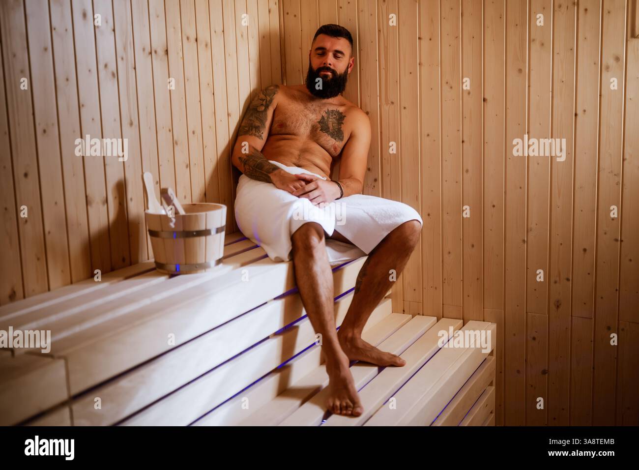 Ein erwachsener Mann lehnt sich gegen die Holzbank einer schwach beleuchteten Sauna, seine Augen geschlossen in friedlicher Glückseligkeit. Stockfoto