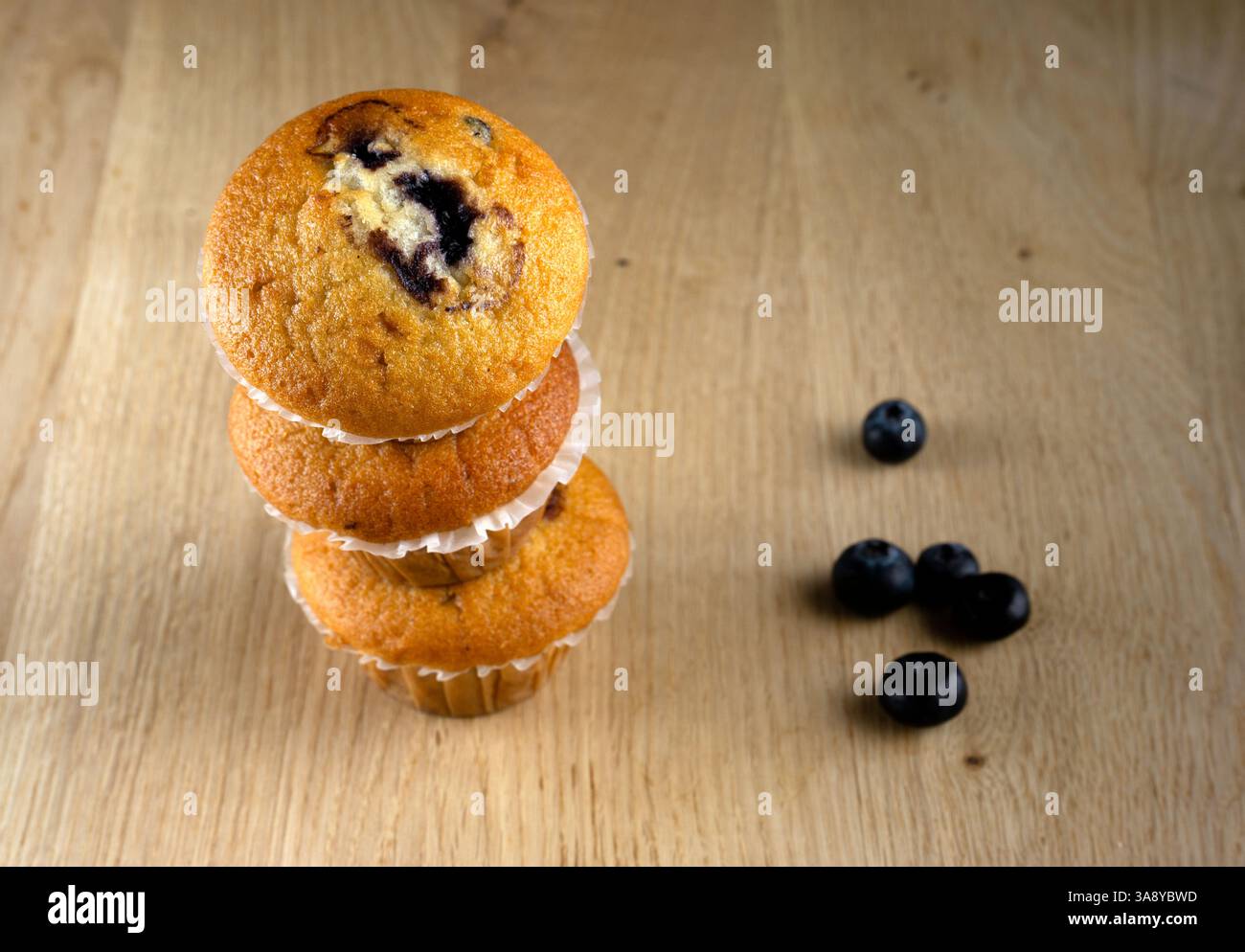 Drei Muffins übereinander gestapelt mit Blaubeeren am Boden. Die Muffins sind mit Heidelbeeren belegt und sitzen auf einem Holztisch Stockfoto