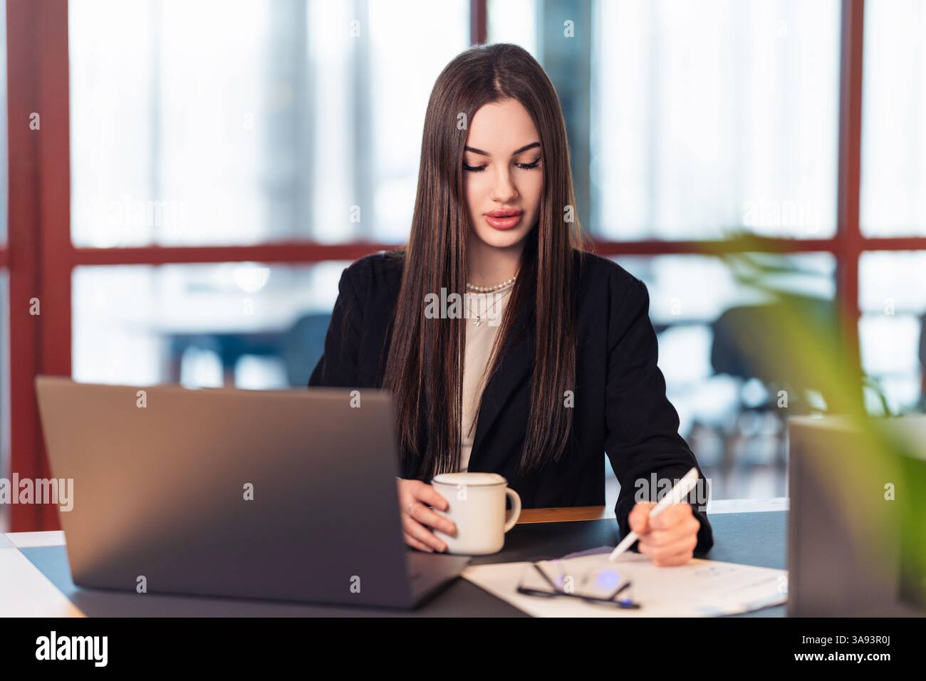 Kaffee, E-Mails, Fristen. Eine Geschäftsfrau tippt auf ihrem Laptop in einem hellen Büro und gleicht Meetings und Projekte aus. Der tägliche Rhythmus des Unternehmens Stockfoto