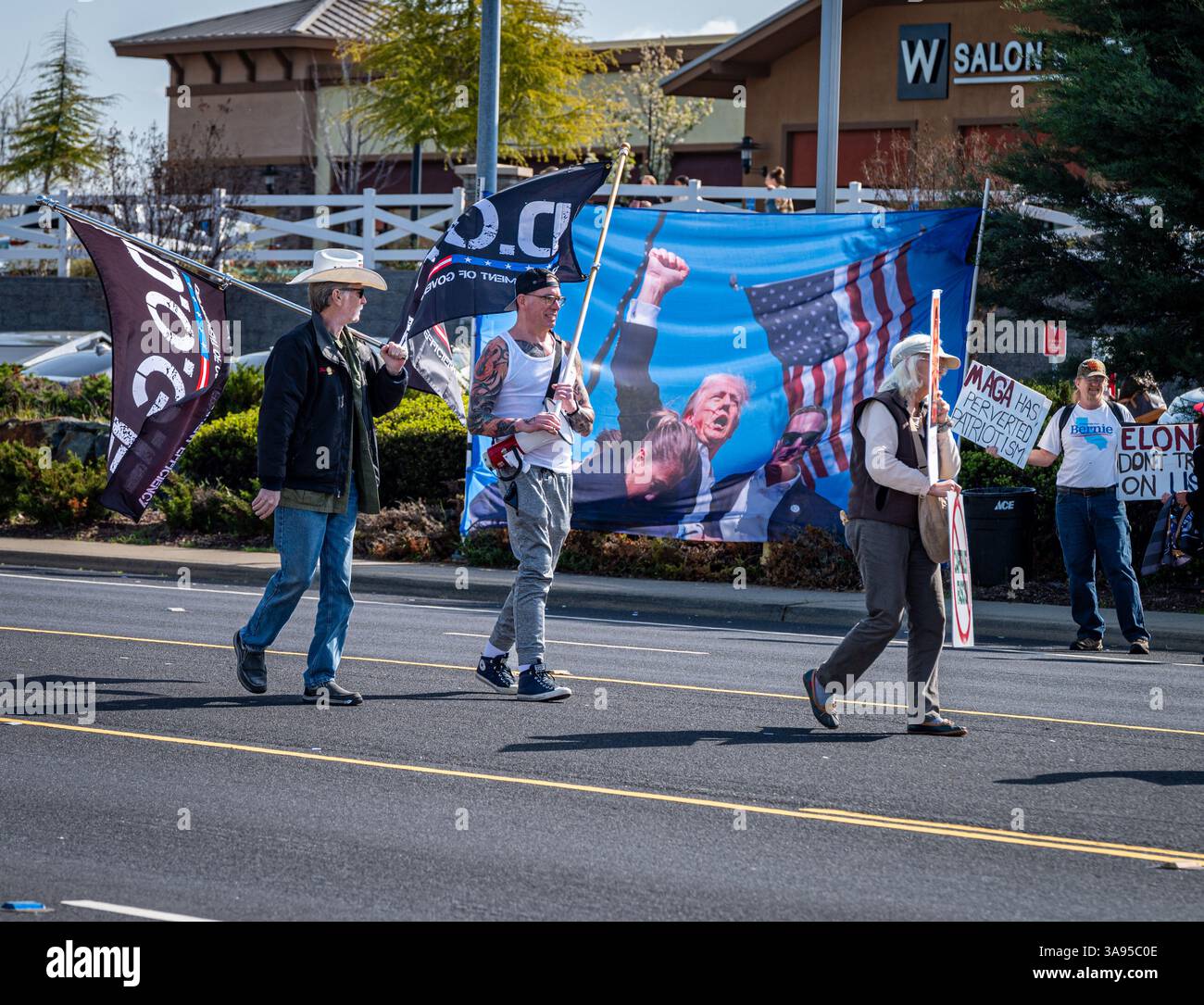 DOGEN-Anhänger gehen an einem Banner eines verletzten Pressers Trump und gegnerischen Aktivisten vorbei, mit einem Schild über MAGA, der Patriotismus und Elon Musk pervertiert. Stockfoto