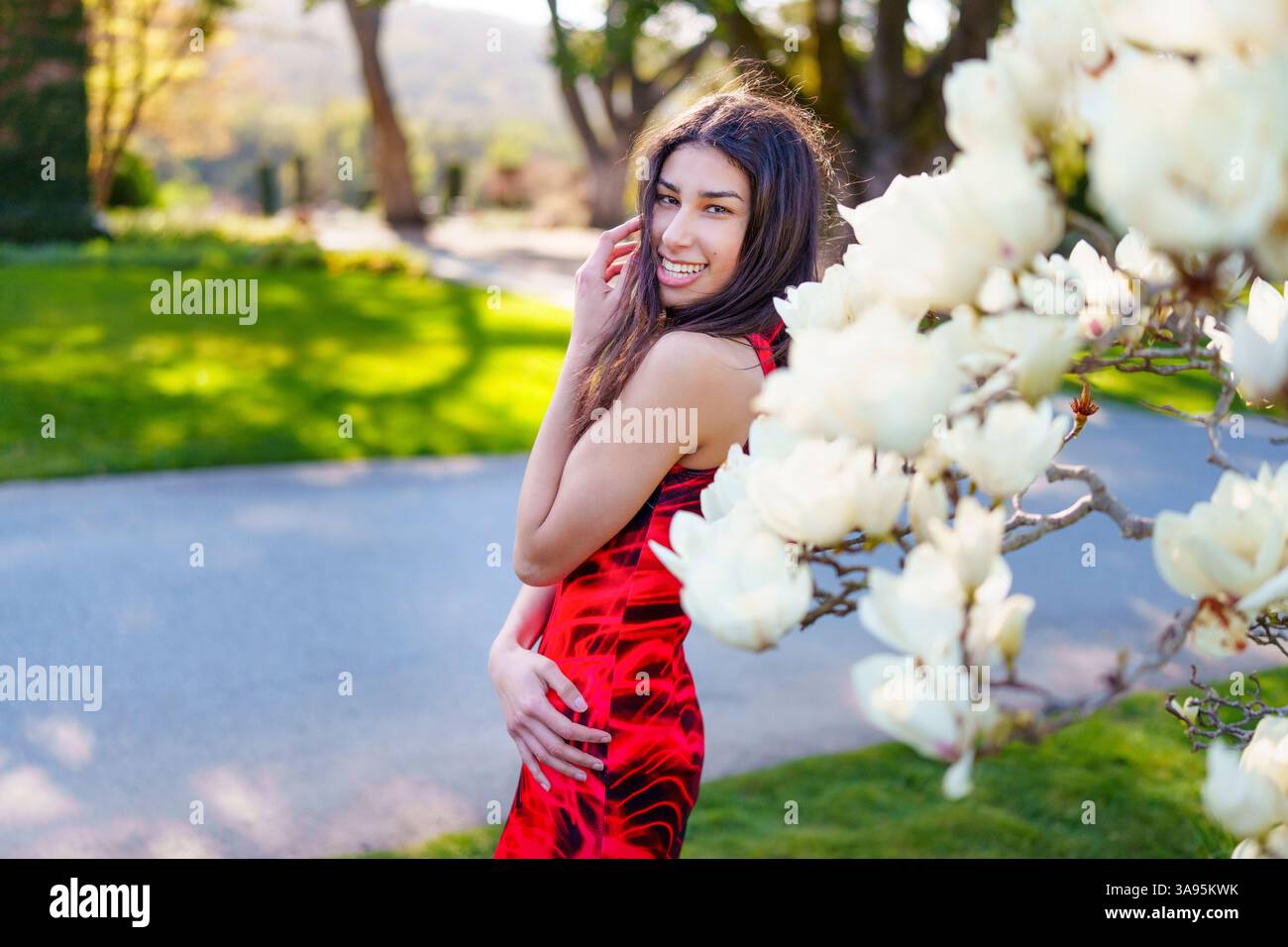 Eine Frau in einem atemberaubenden roten Kleid genießt einen schönen Frühlingstag umgeben von blühenden Magnolienblüten Stockfoto