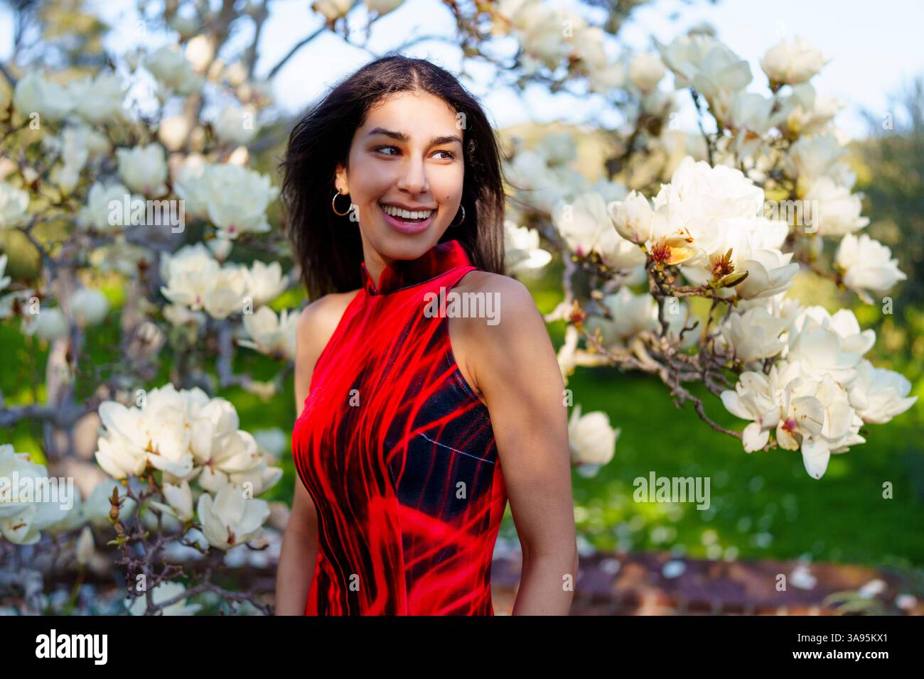 Eine Frau in einem atemberaubenden roten Kleid genießt einen schönen Frühlingstag umgeben von blühenden Magnolienblüten Stockfoto