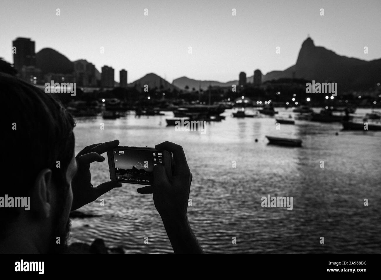 Ein Mann fotografiert den Corcovado und die Guanabara-Bucht in der Abenddämmerung in Urca – Rio de Janeiro, Brasilien Stockfoto