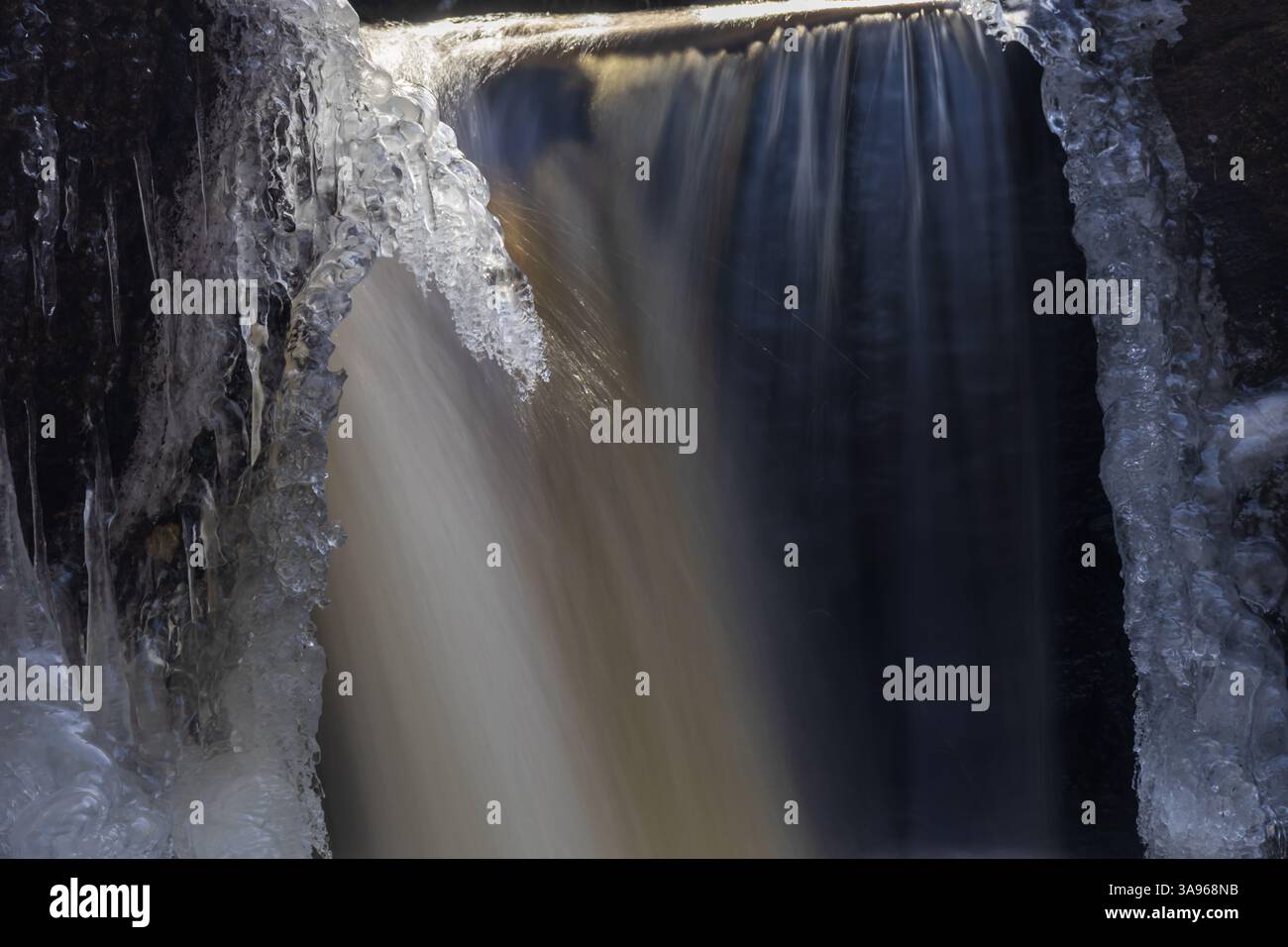 Nahaufnahme eines gefrorenen Wasserfalls mit nebligem fließendem Wasser, kurzen Eiszapfen, die von Felsen hängen, und Eisformationen, die die Steinoberfläche bedecken Stockfoto