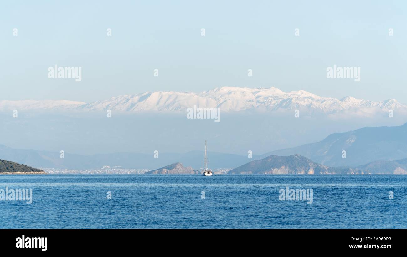 Ein Segelboot schwimmt auf dem Mittelmeer in Fethiye, Türkei, mit einer atemberaubenden Kulisse aus schneebedeckten Bergen und einem klaren blauen Himmel, was eine ruhige Mutter schafft Stockfoto