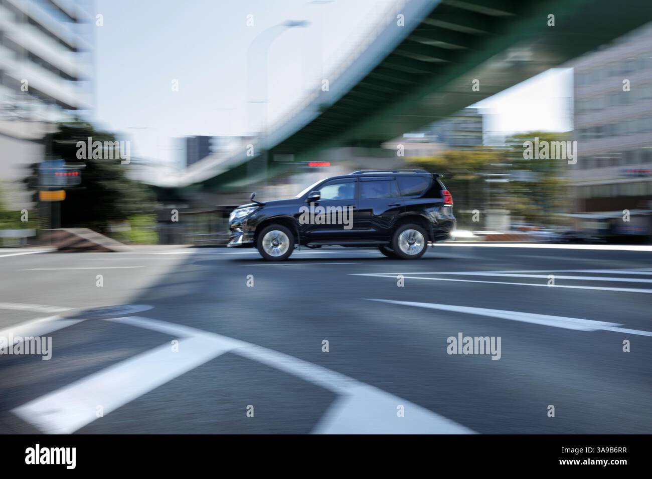 Der schwarze SUV fährt durch eine städtische Kreuzung mit Gebäuden und einer Überführung. Bewegungsunschärfe betonen die Fahrzeugbewegung und die geschäftige Stadt. Stockfoto