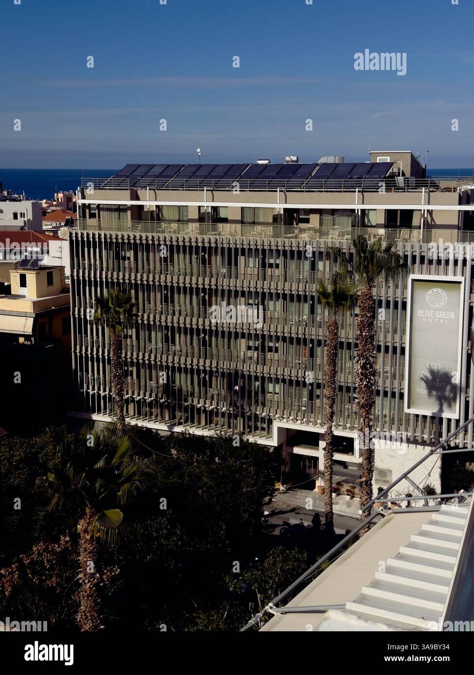 Blick von oben auf das Green Olive Hotel mit einem Schild mit der Aufschrift „Eco, Friendly and Smart Hospitality“, Palmen und im Hintergrund die Stadt Heraklion Stockfoto