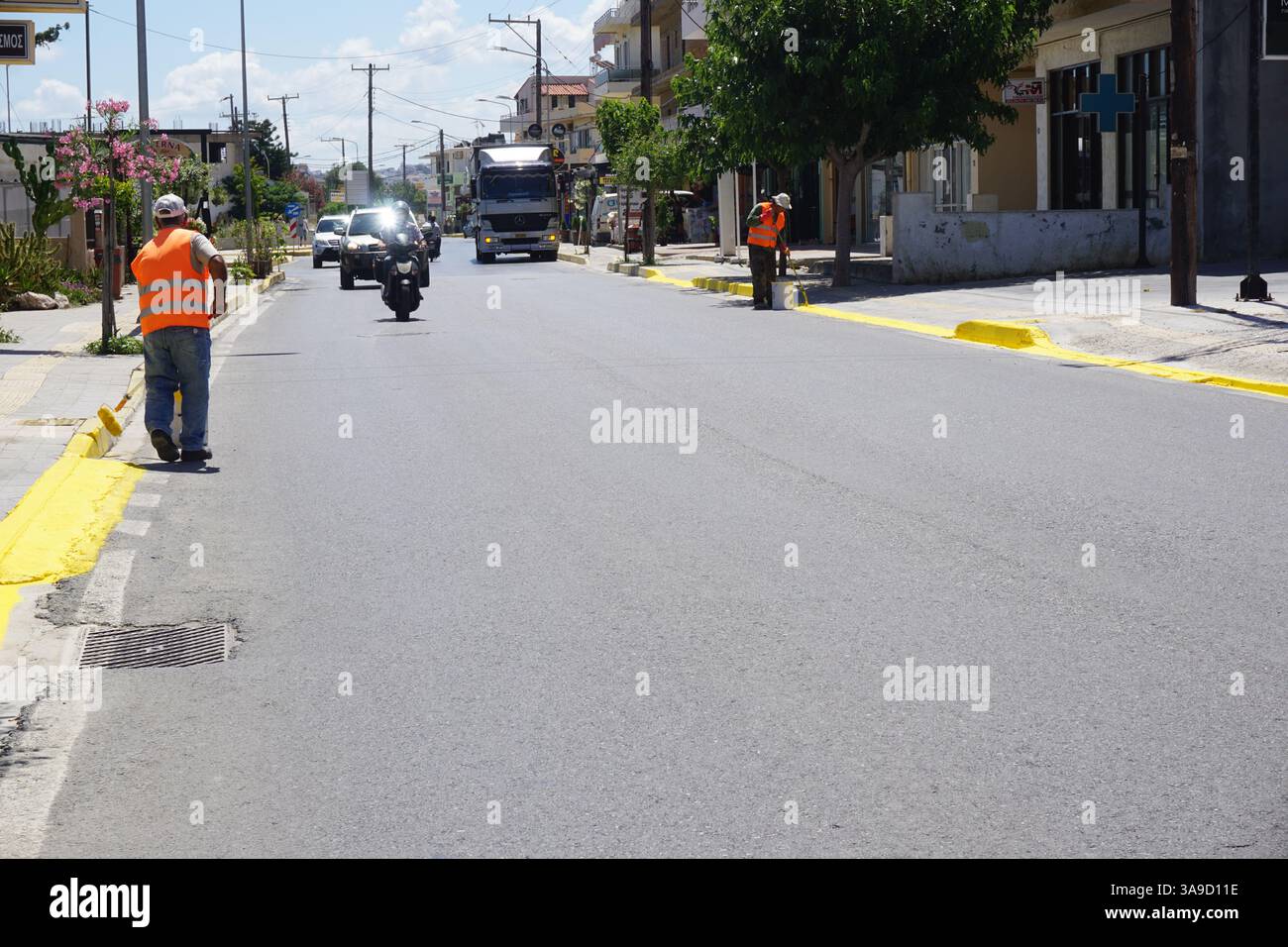 Zwei Arbeiter in fluoreszierenden Westen halten Pinsel und lackieren die Bordsteinkanten mit gelber Farbe auf einer Straße Stockfoto