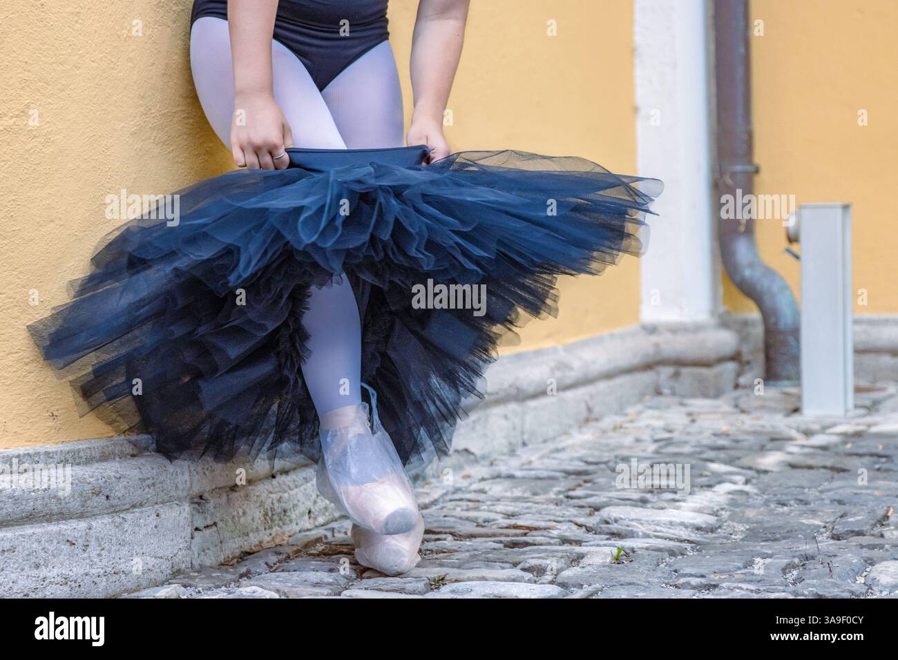 Schließen Sie junge weibliche Ballerina, die sich verkleidet und schwarzes Tutu an der gelben Gebäudewand anzieht. Ballett-Tänzer nimmt den Rock. Punkte Stockfoto