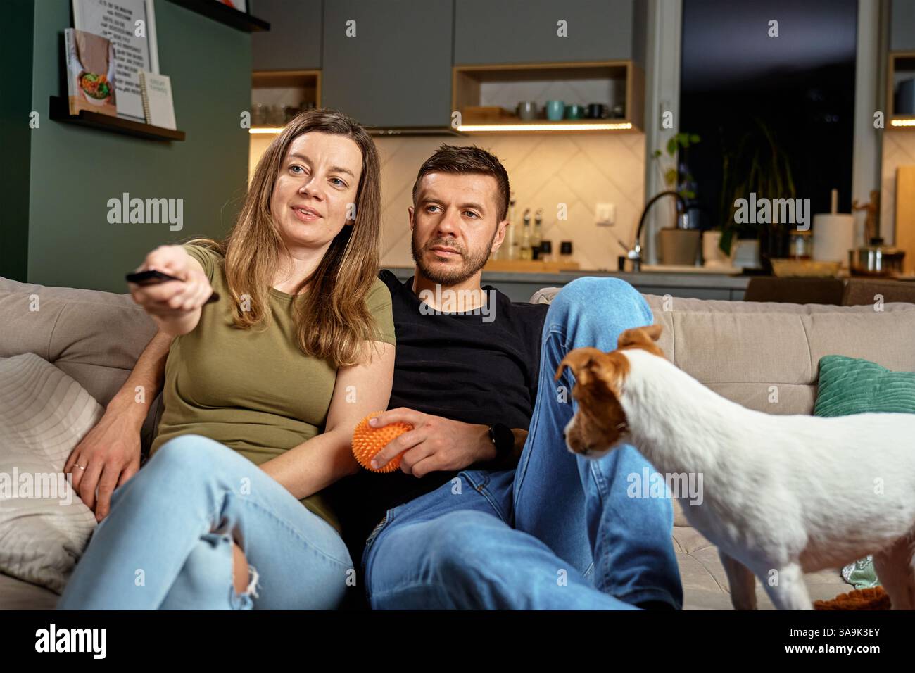Mann und Frau sitzen auf Couch mit Fernbedienung und Hund. Paare schauen zusammen zu Hause fern. Konzept der häuslichen Entspannung, Gesellschaft und gemütliches häusliches Leben Stockfoto