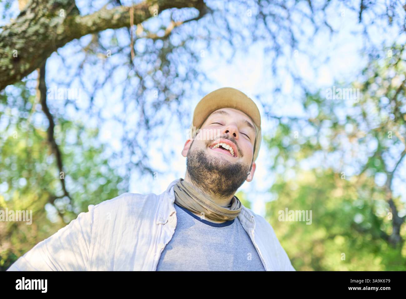 Porträt eines jungen Mannes, der mit einem Ausdruck von Glück und Wohlbefinden im Freien, in der Natur lächelt; der Hintergrund mit dem Kopierraum ist Himmel und Baum Stockfoto