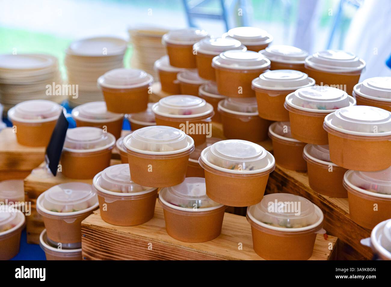 Mehrere Lebensmittelbehälter aus braunem Papier mit Kunststoffdeckeln, die ordentlich auf Holzständern angeordnet sind und für die Verteilung bei einer Veranstaltung mit Catering bereit sind. Stockfoto