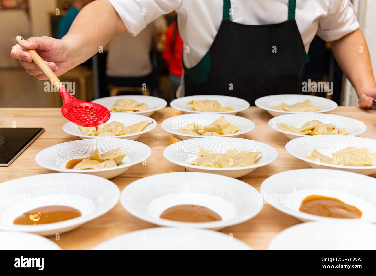 Ein Koch in einer schwarzen Schürze serviert frisch gekochte Ravioli mit Sauce zu mehreren weißen Gerichten, die in einem Restaurant serviert werden. Stockfoto