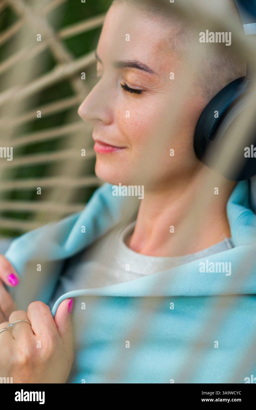 Junge weibliche Krebspatientin, die sich im Hängestuhl auf der Gartenterrasse entspannt und Musik hört. Geistiges Wohlbefinden während der Krankheit. Stockfoto