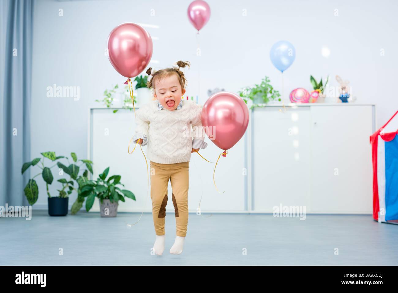 Niedliches 3-jähriges Mädchen mit Down-Syndrom feiert ihren Geburtstag in einem mit Ballons dekorierten Spielzimmer. Kindergeburtstag. Stockfoto