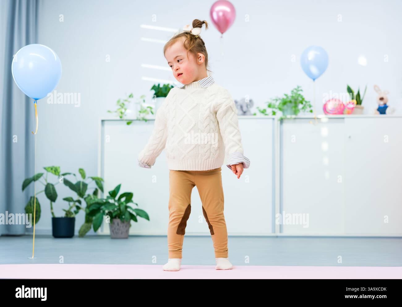 Niedliches 3-jähriges Mädchen mit Down-Syndrom feiert ihren Geburtstag in einem mit Ballons dekorierten Spielzimmer. Kindergeburtstag. Stockfoto