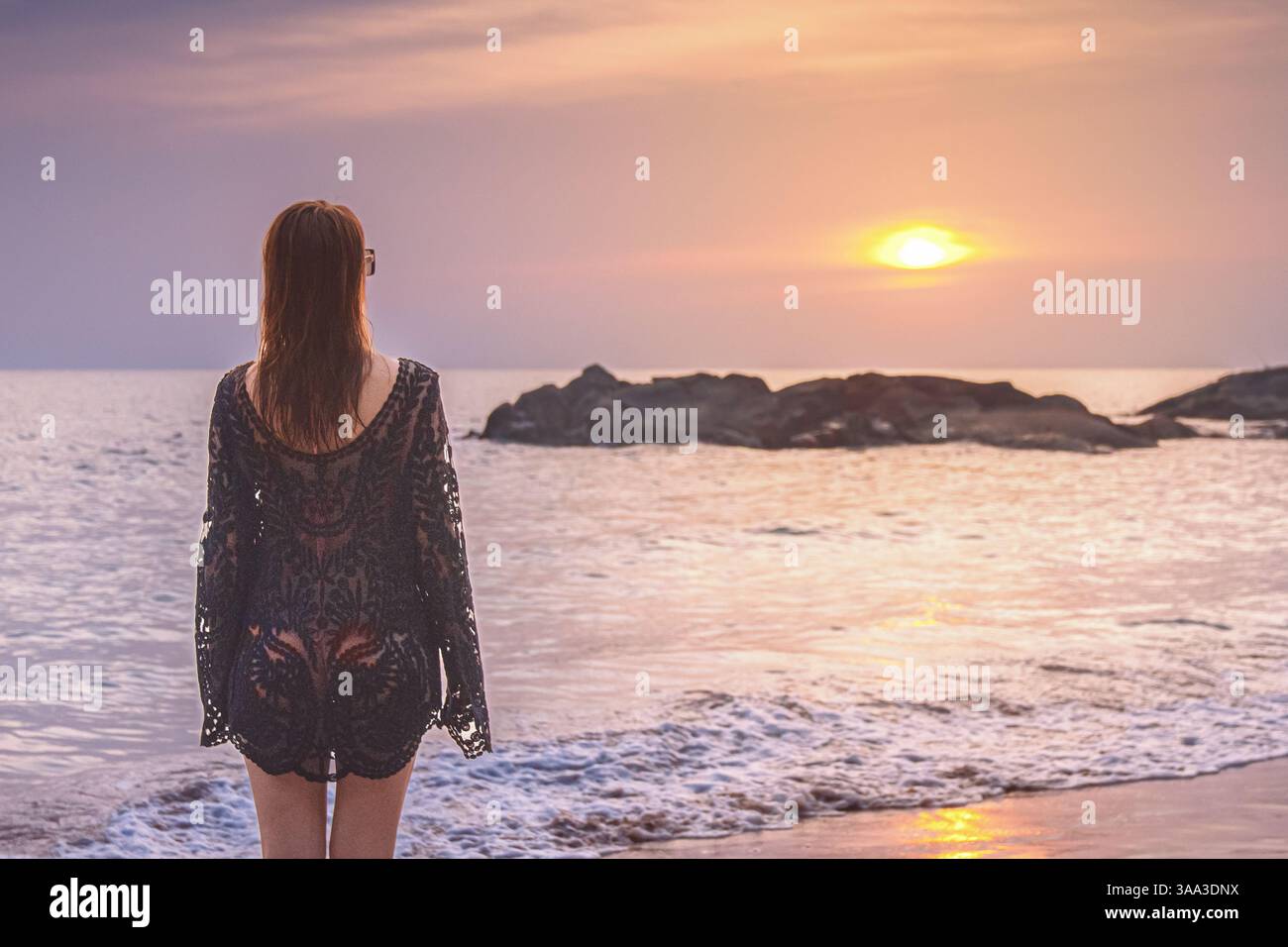 Alleinreisen. Eine Frau in einem schwarzen cape schaut auf den Sonnenuntergang am Strand in Khao Lak, Thailand Stockfoto