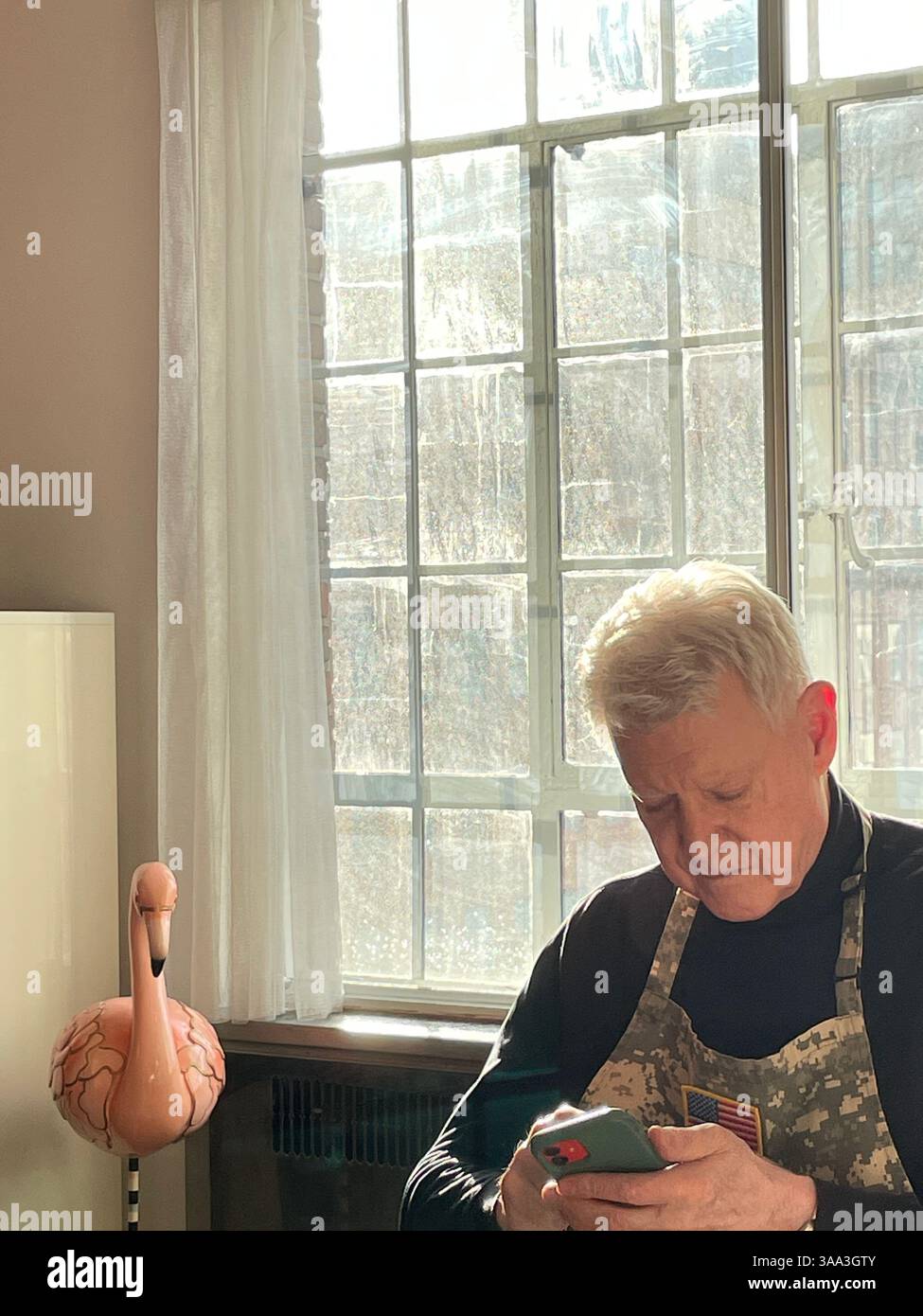 Senior Mann in seiner Wohnung, der mit seinem iPhone spielt, während er die schmutzigen Fenster ignoriert, 2025, New York City, USA - Smartphone-aufgenommenes Stockfoto