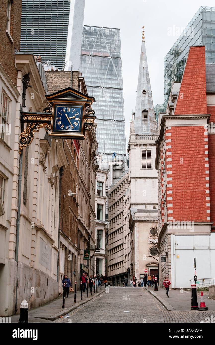 Ein hoher Kirchturm erhebt sich zwischen modernen Londoner Türmen, eine enge Kopfsteinpflasterstraße mit Menschen, die unter einer alten Straßenuhr spazieren gehen - 04.01.2024, Großbritannien, Londo Stockfoto