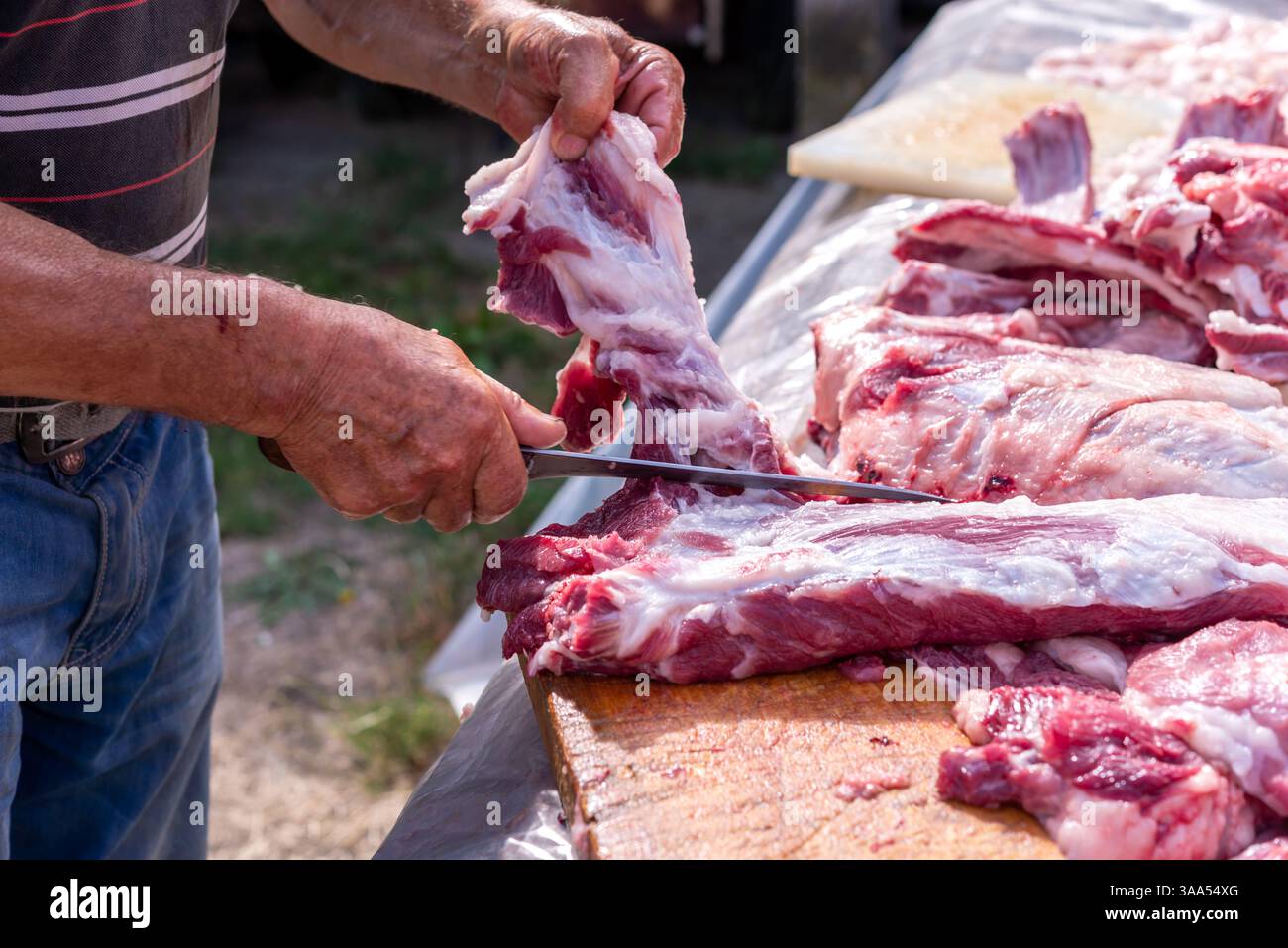 Die Hände eines erwachsenen Mannes schneiden rohes Schweinefleisch mit einem Messer auf einem hölzernen Schneidebrett draußen. Stockfoto
