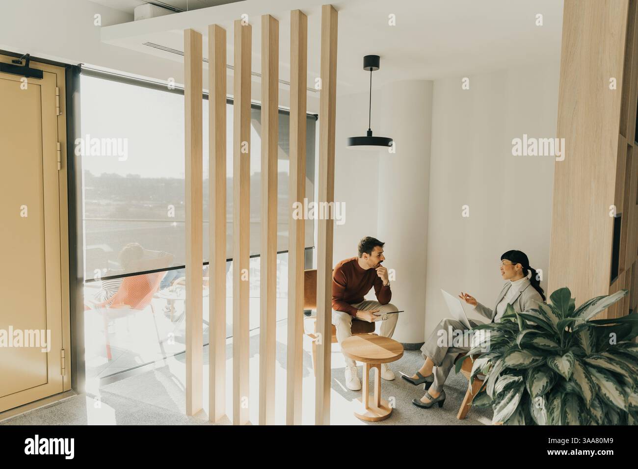 Zwei Personen in zeitgenössischer Kleidung unterhalten sich in einer hellen Lounge mit Holzakzenten und Grün. Das Sonnenlicht wirft sanfte Schatten, Anzeige Stockfoto