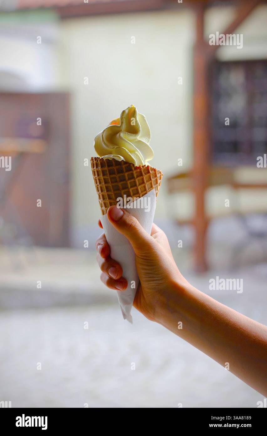 Die Hand des kaukasischen Jungen hält Vanilleeiskegel in einer weißen Serviette, im Hintergrund ein verschwommenes Gartenrestaurant. Selektiver Fokus, Kopierraum Stockfoto