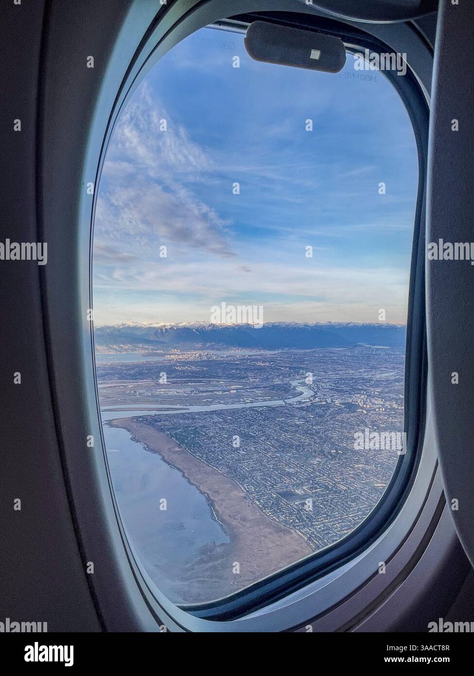 Ein Blick auf die Stadt und die Küste durch ein Flugzeugfenster mit blauem Himmel und fernen Bergen im Hintergrund. - Smartphone-aufgenommenes Stockfoto