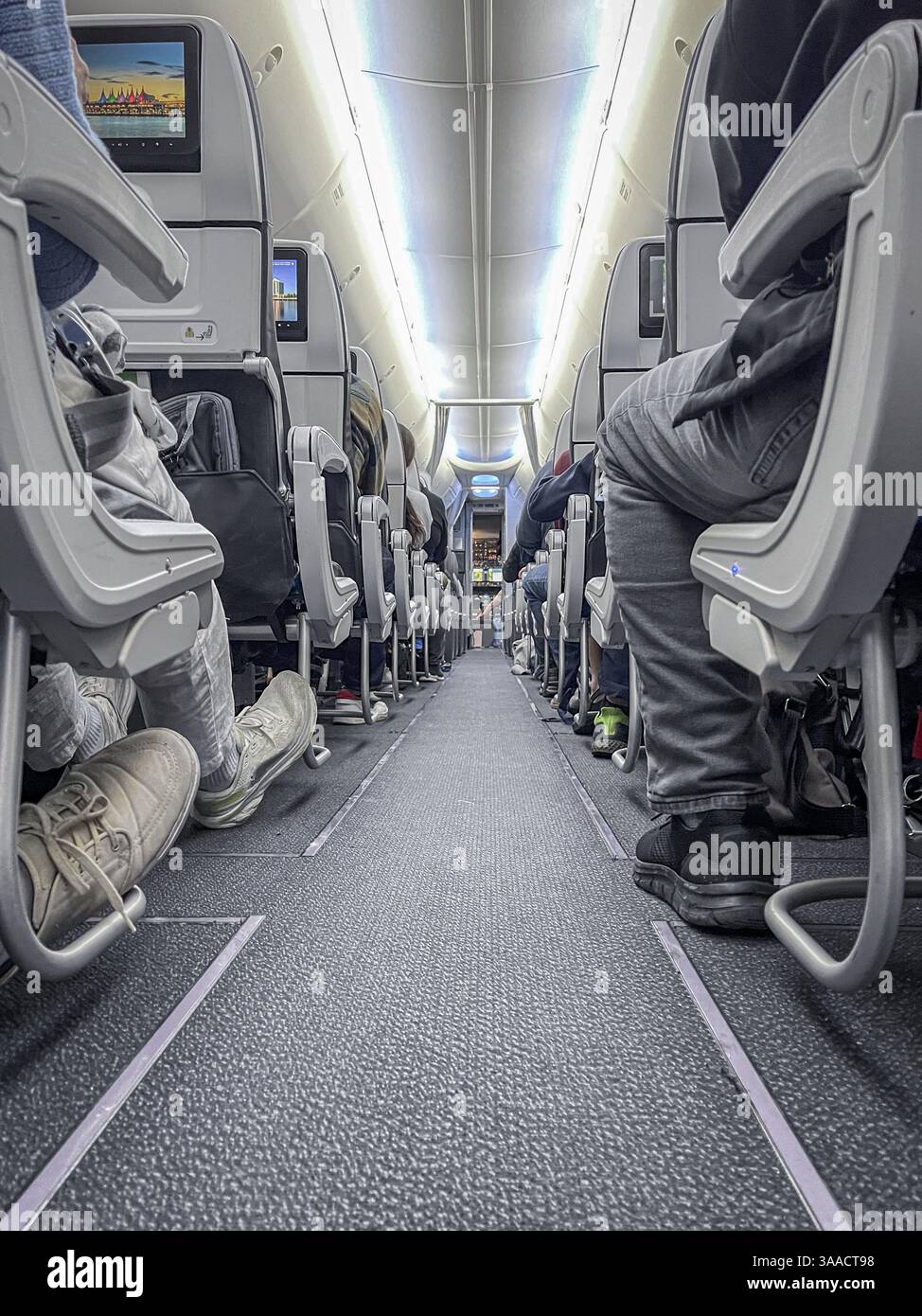 Innenansicht der Flugzeugkabine mit Fluggästen, die mit Blick auf den Gang in Richtung Cockpit sitzen, beleuchtet durch Kabinenbeleuchtung. - Smartphone-aufgenommenes Stockfoto