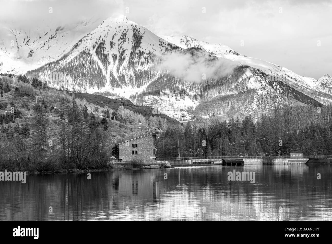 Ein Steinhaus an einem ruhigen See, umgeben von alpinen Bäumen, mit majestätischen schneebedeckten Bergen im Hintergrund unter einem bewölkten Himmel. Stockfoto