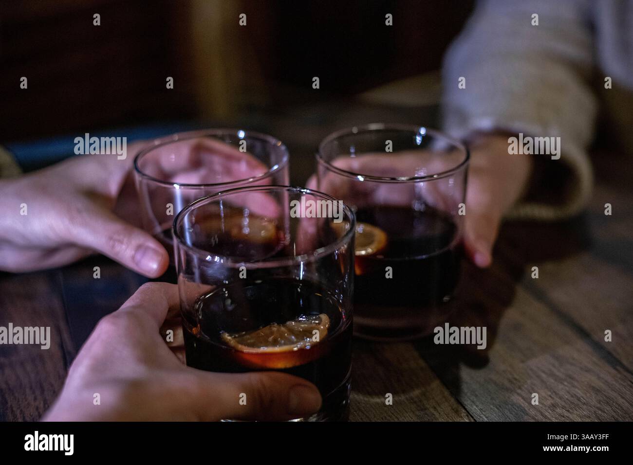 Drei Freunde feiern einen Geburtstag, indem sie drei Gläser alkoholischer Getränke mit Kirschen in einem Club im gestreuten Licht anschlagen Stockfoto