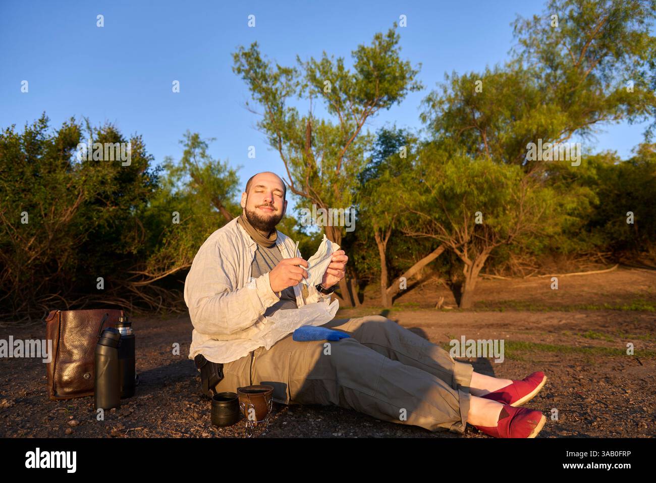Der Reisende sitzt bei Sonnenaufgang am Ufer eines Baches und empfängt die ersten Sonnenstrahlen mit positivem Ausdruck, während er sich darauf vorbereitet, Mate zu trinken, ein Typi Stockfoto