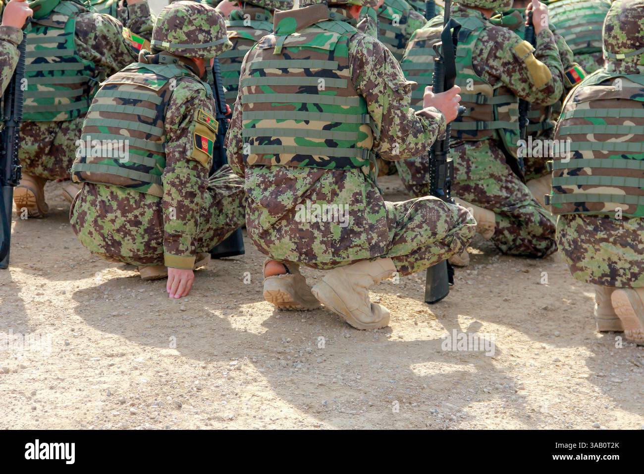 Eine Gruppe Soldaten in getarnten Uniformen kniet draußen, ausgestattet mit taktischer Ausrüstung. Sie scheinen bereit für Trainingsübungen in einem trockenen, draußen Stockfoto