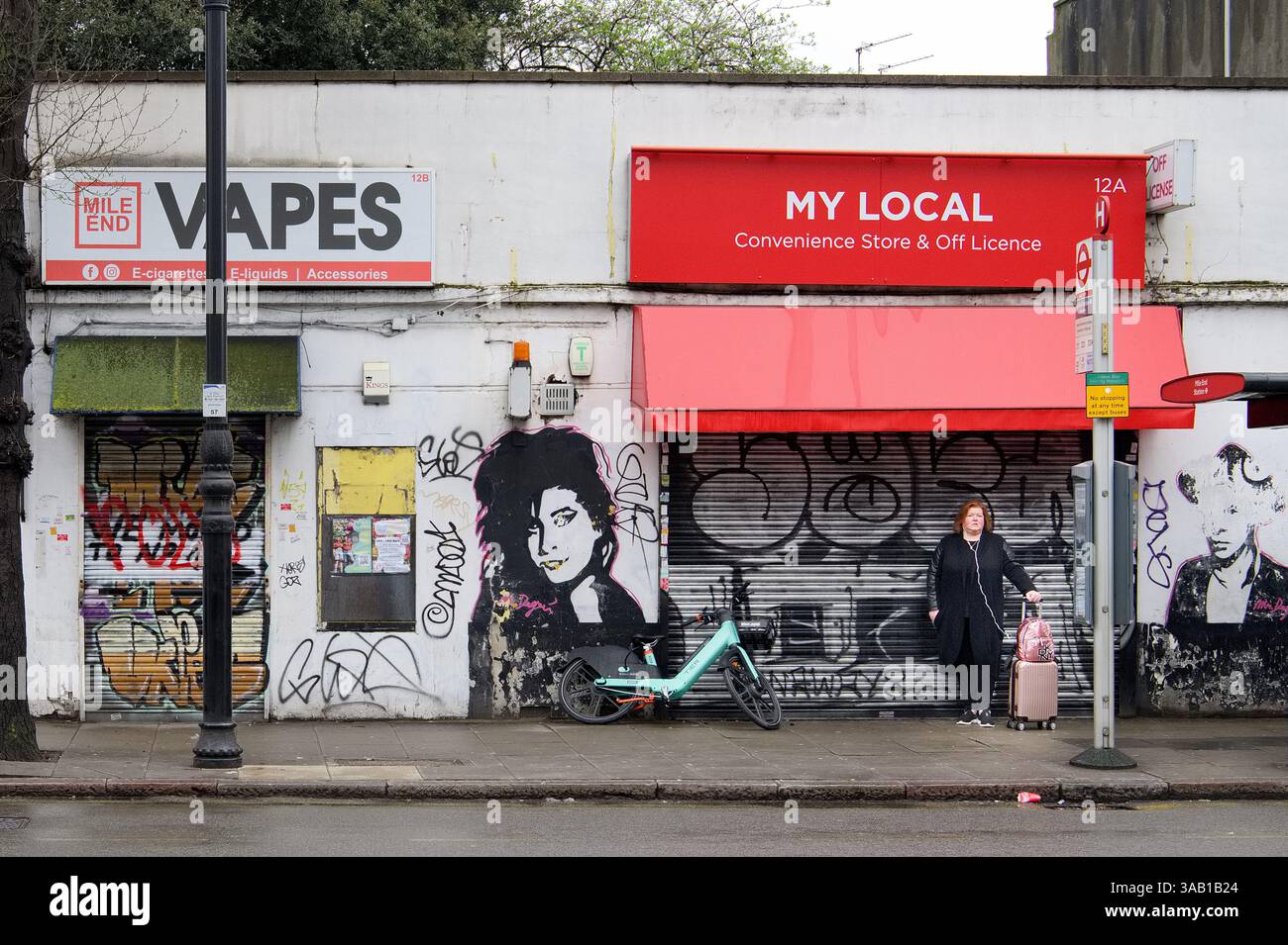 Frau mit rosafarbenem Koffer an der Bushaltestelle, umgeben von Graffiti und einem schwarz-weißen Wandgemälde von Amy Winehouse, Grove Road, Mile End, London Stockfoto