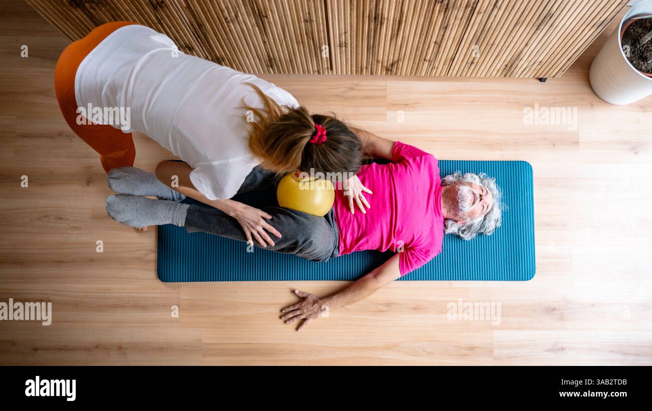 Eine Frau in orangefarbenen Leggings führt einen älteren Mann in einem rosafarbenen Hemd durch eine Kernübung mit einem gelben Ball, der in einer Fitnesssitzung auf einer blauen Matte liegt. Stockfoto