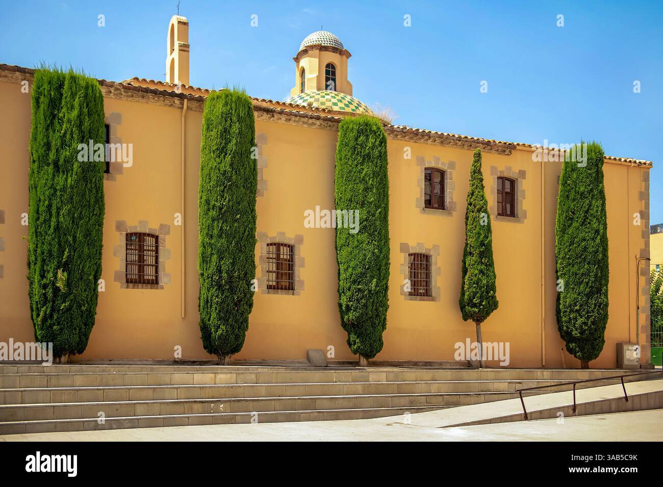 Hohe Zypressen, die symmetrisch entlang einer alten gelben Stuckwand mit Gitterfenstern und einer bunten gekachelten Kuppel unter einem klaren blauen Himmel stehen Stockfoto